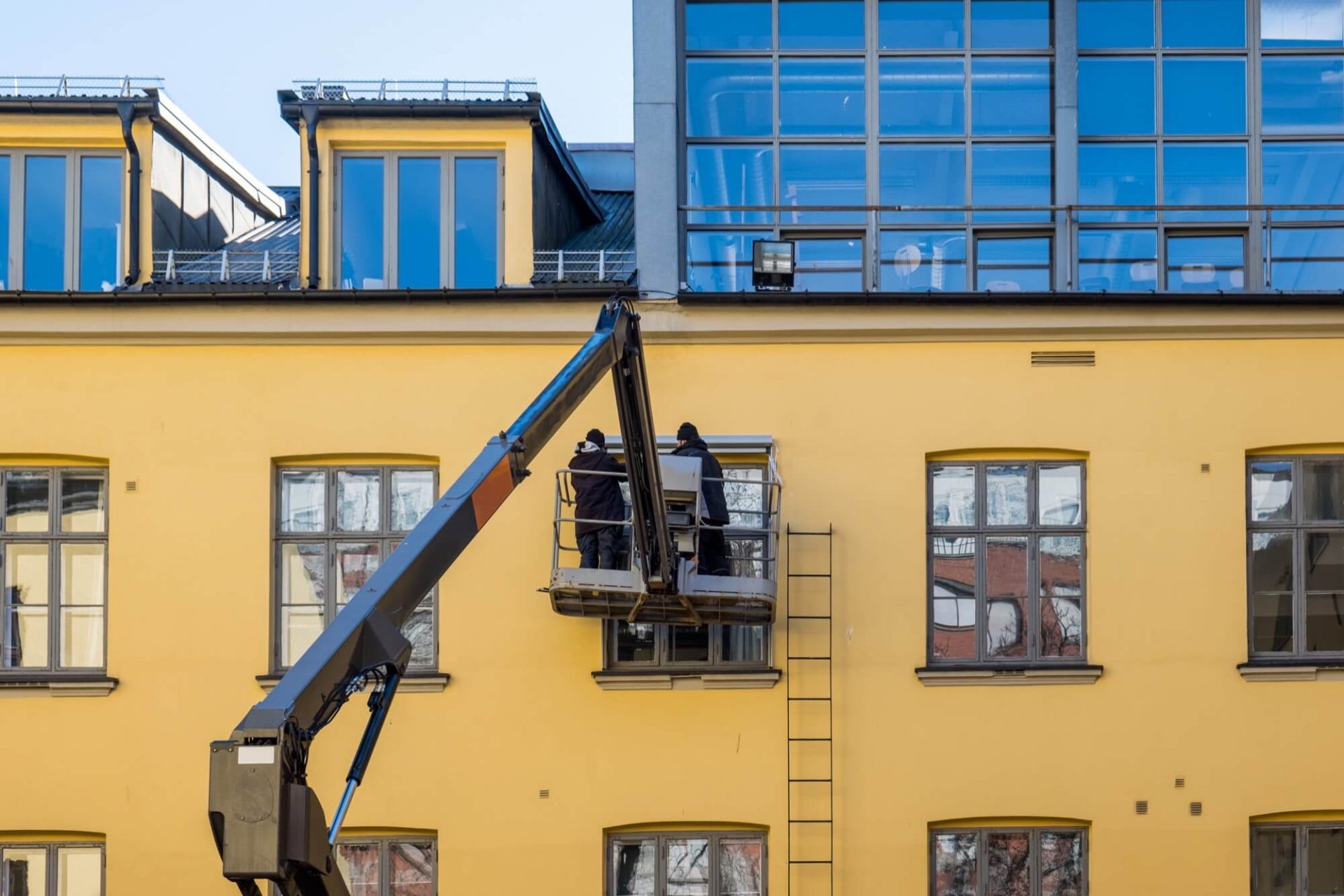 På en gul byggnad med stora fönster ses arbetare i en skylift som utför fasadarbeten